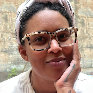 A woman with dark curly hair, wearing a white headscarf and tortoiseshell glasses, touching her face gently, outdoors with a stone wall in the background.