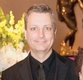 A man with short gray hair wearing a black suit, black shirt, and black tie, smiling in a setting with yellow flowers and a gold decorative sculpture in the background.