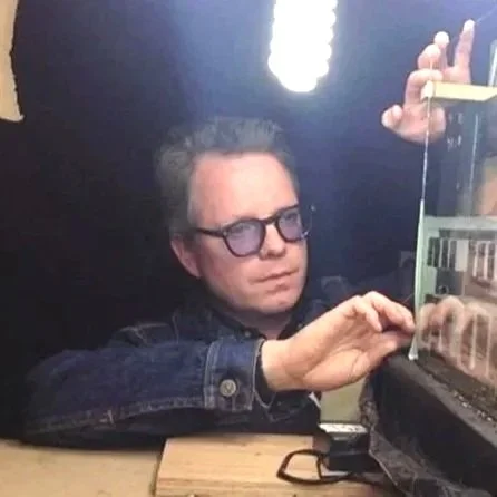 A man with glasses working on or inspecting a glass display case in a dimly lit room with bright ceiling lights.