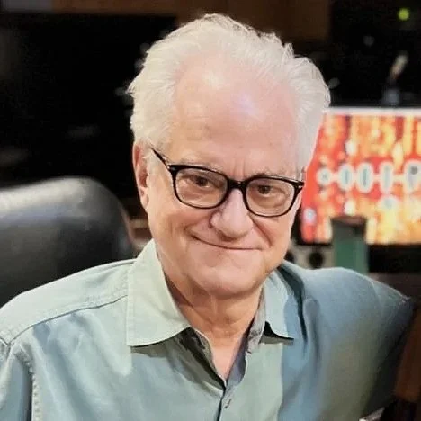 An elderly man with white hair, glasses, and a light green shirt smiling in a recording studio.