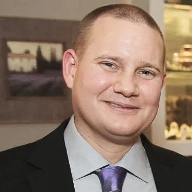 A smiling man wearing a black suit, light blue shirt, and purple tie, in an indoor setting with artwork and shelves in the background.