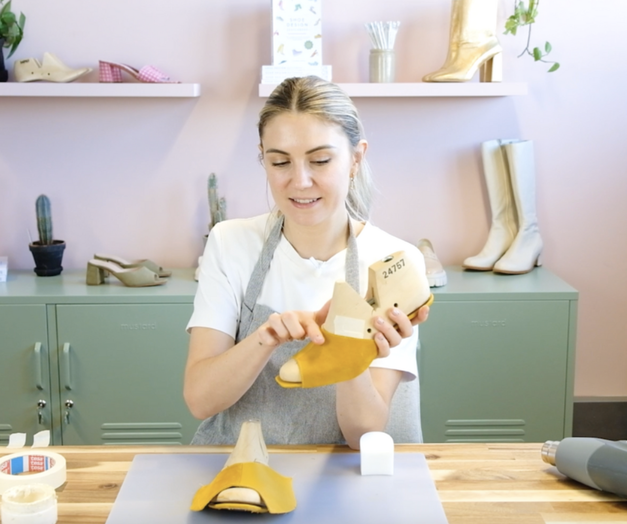 A woman in a white shirt and gray apron is inspecting a yellow high-heeled shoe she is holding, in a workshop with various shoes and small potted cacti in the background.