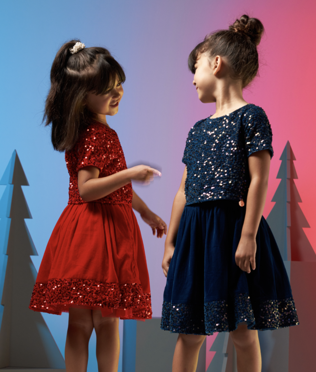 Two young girls wearing sparkly dresses standing face to face indoors with colorful lighting and decorated trees in the background.