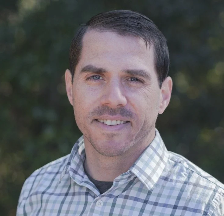 John Karabees, LPC, smiling and wearing a plaid button-down shirt, posing in a three-quarter pose in front of a green leafy background.