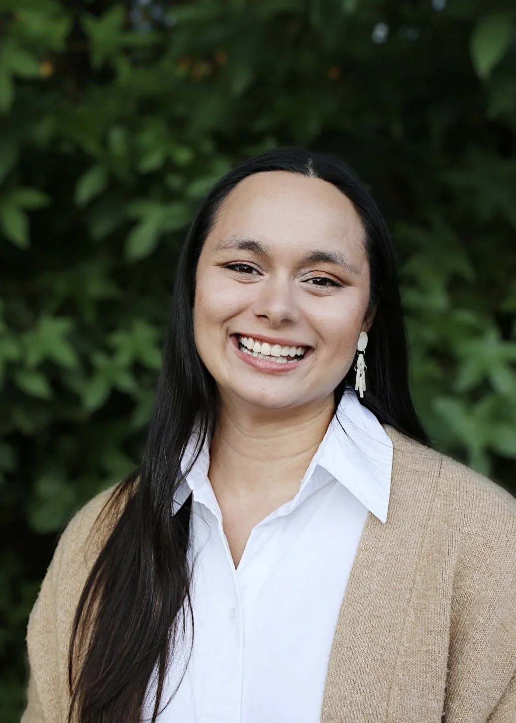 Meredith Yuhas Gawai, LPC, smiling with long dark hair, wearing a white blouse and tan cardigan, posing in a three-quarter pose in front of a green leafy background.
