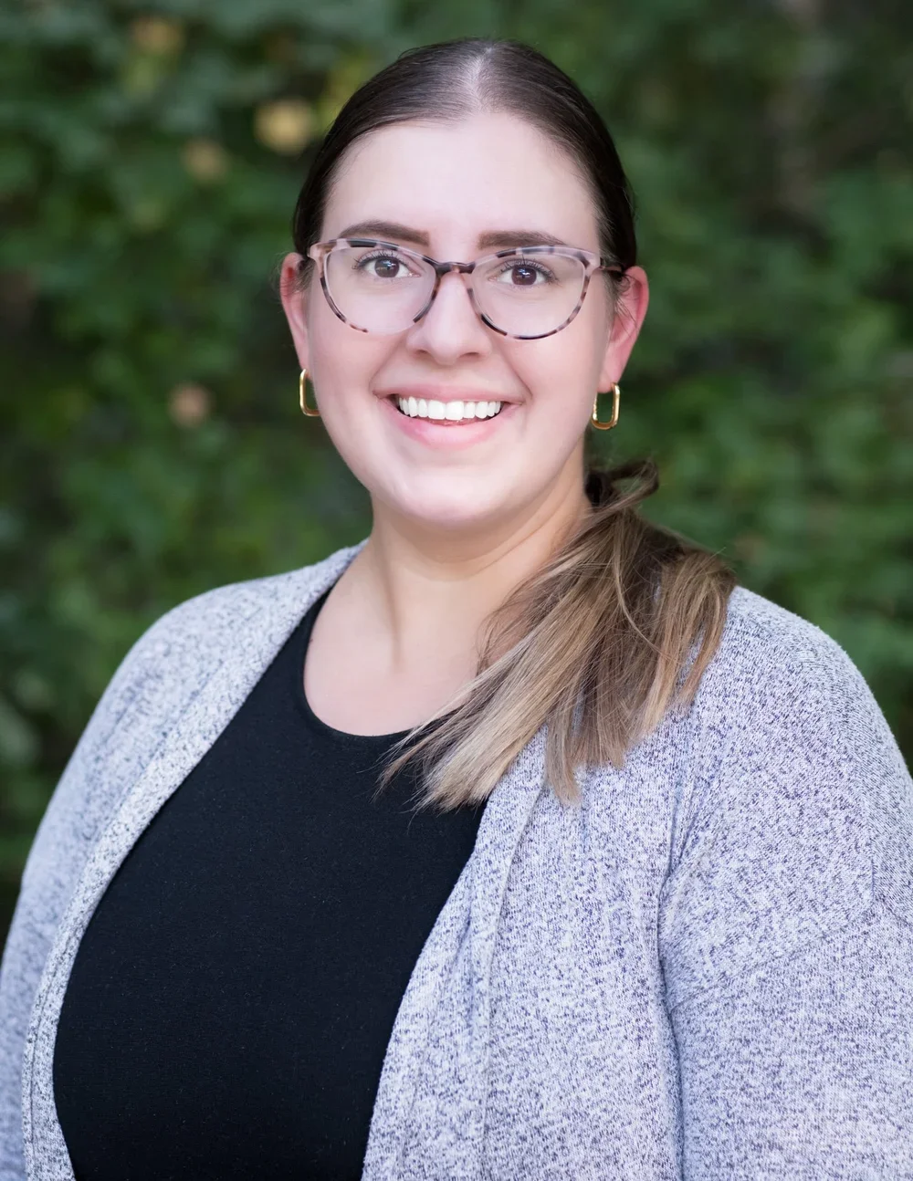 Melissa Colleton, LMSW, smiling and wearing glasses, a black top, and a light cardigan, posing in a three-quarter pose in front of a green leafy background.
