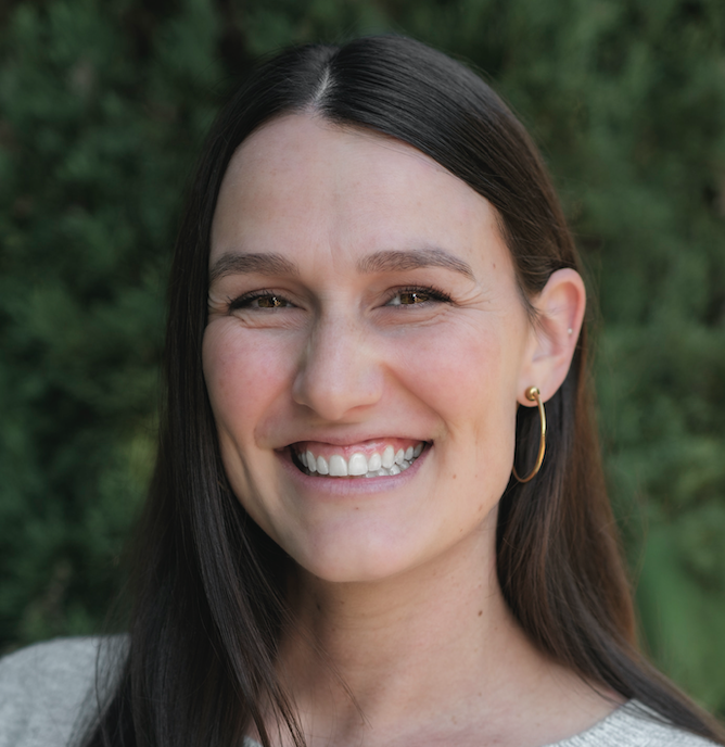 Katherine Cristan, LPC - Smiling woman with long dark hair, wearing gold hoop earrings and a light gray sweater, photographed outdoors in front of green foliage