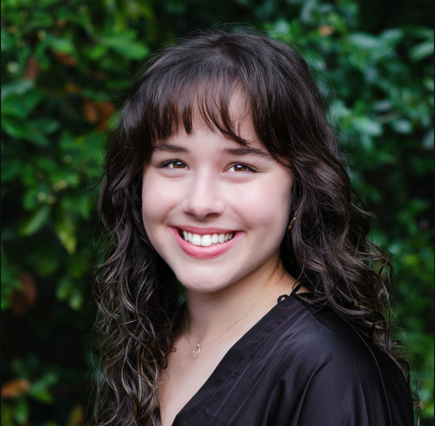 Sarah Mulroney, LPC-A, smiling with shoulder-length dark hair, wearing a black top, posing in a three-quarter pose in front of a green leafy background.