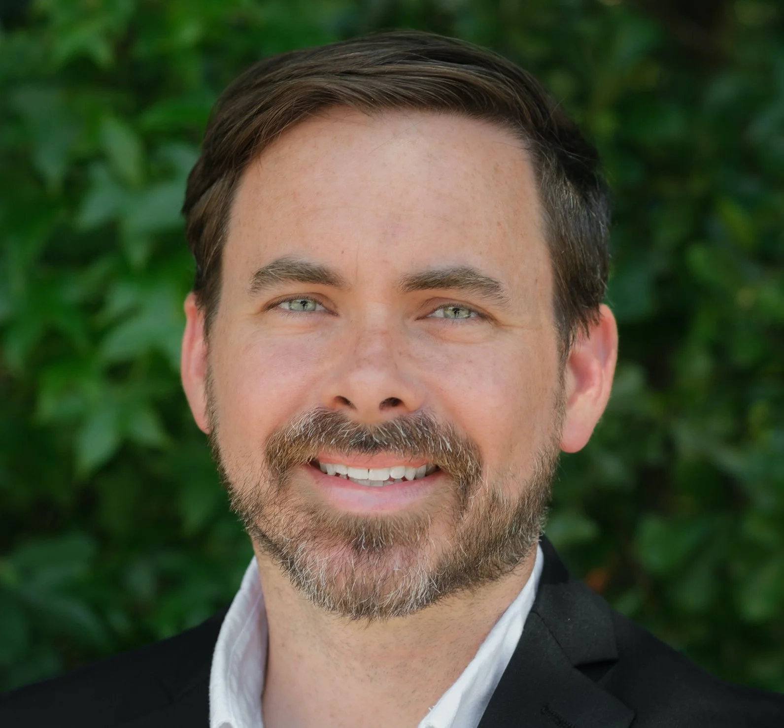 Chad Cooper, LPC-A, smiling with short dark hair and facial hair, wearing a white shirt and dark blazer, posing in a three-quarter pose in front of a green leafy background.