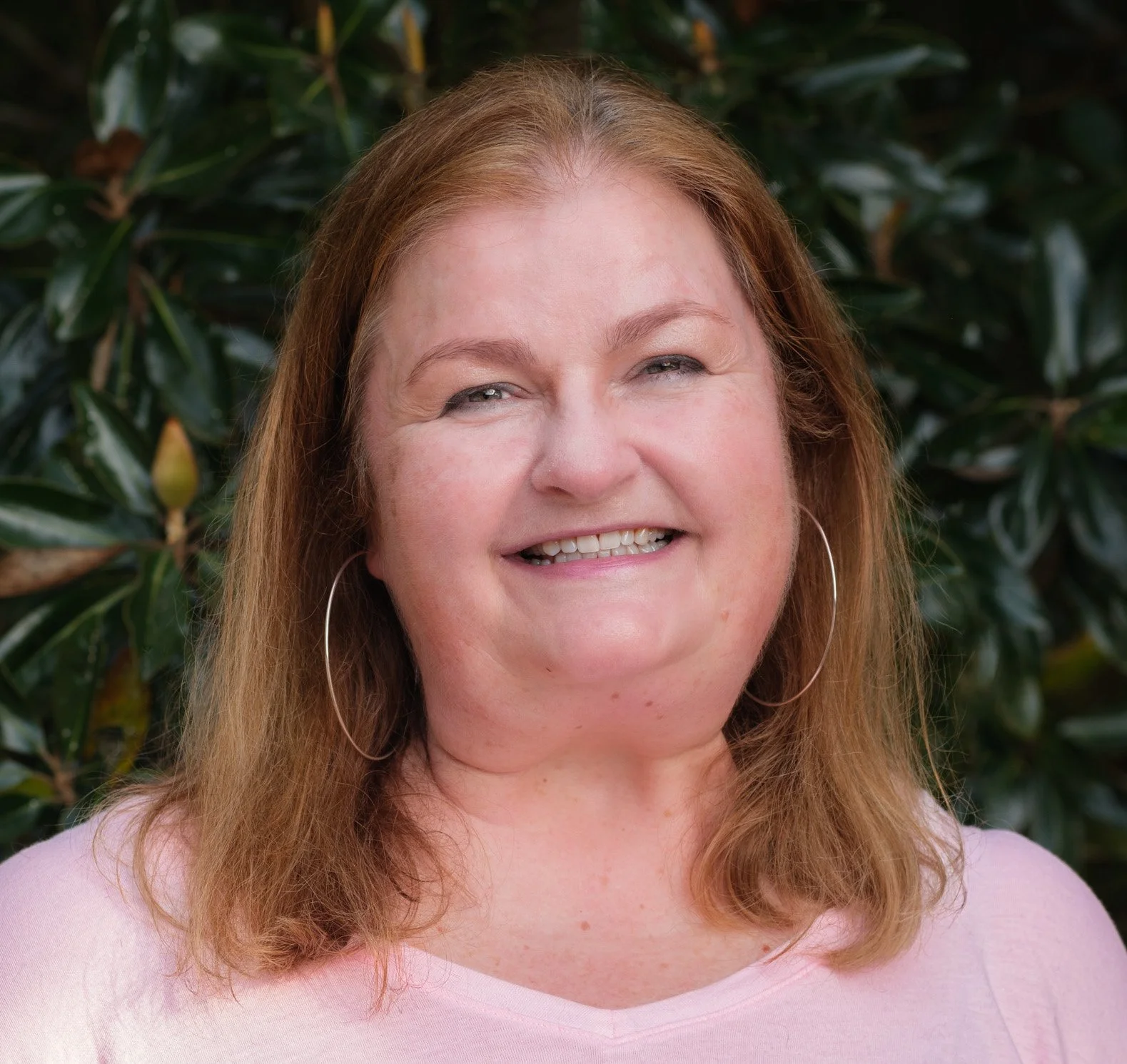 Amy Ashland, LPC, smiling with shoulder-length light brown hair, wearing a light pink top, posing in a three-quarter pose in front of a green leafy background.