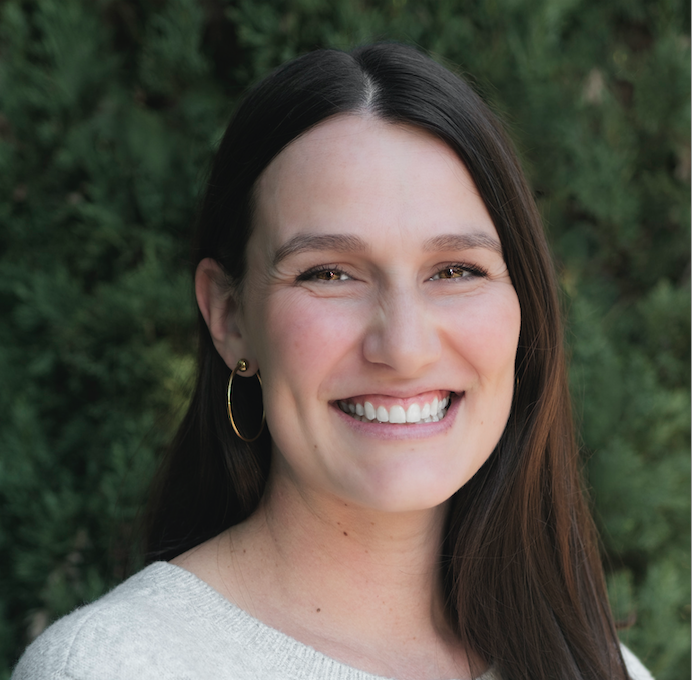 Katherine Cristan, LPC - Smiling woman with long dark hair, wearing gold hoop earrings and a light gray sweater, photographed outdoors in front of green foliage