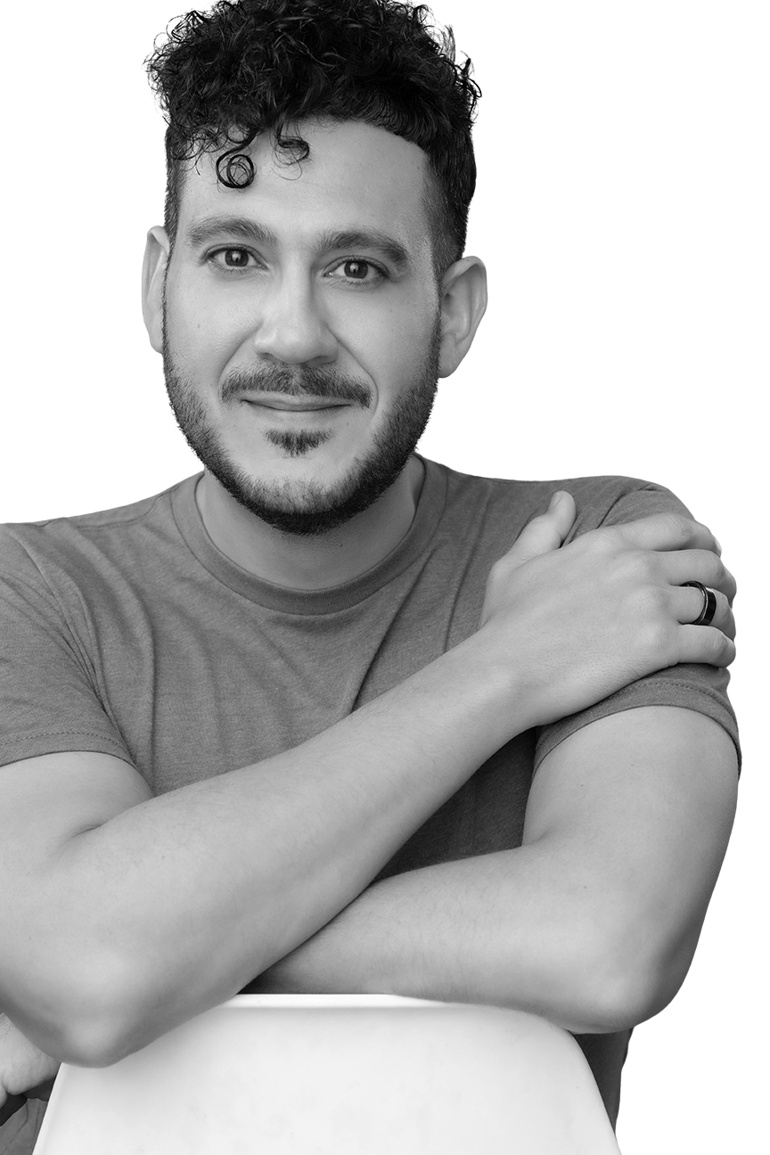 A young man with curly hair and a beard, smiling at the camera, wearing a casual t-shirt, with his arms crossed over an office chair, in black and white.