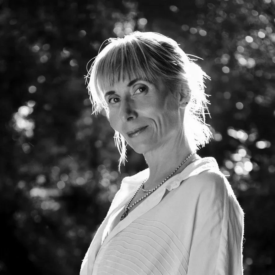 A black-and-white photo of an older woman with short hair, wearing a light-colored top and a necklace, smiling softly outdoors with blurred trees in the background.