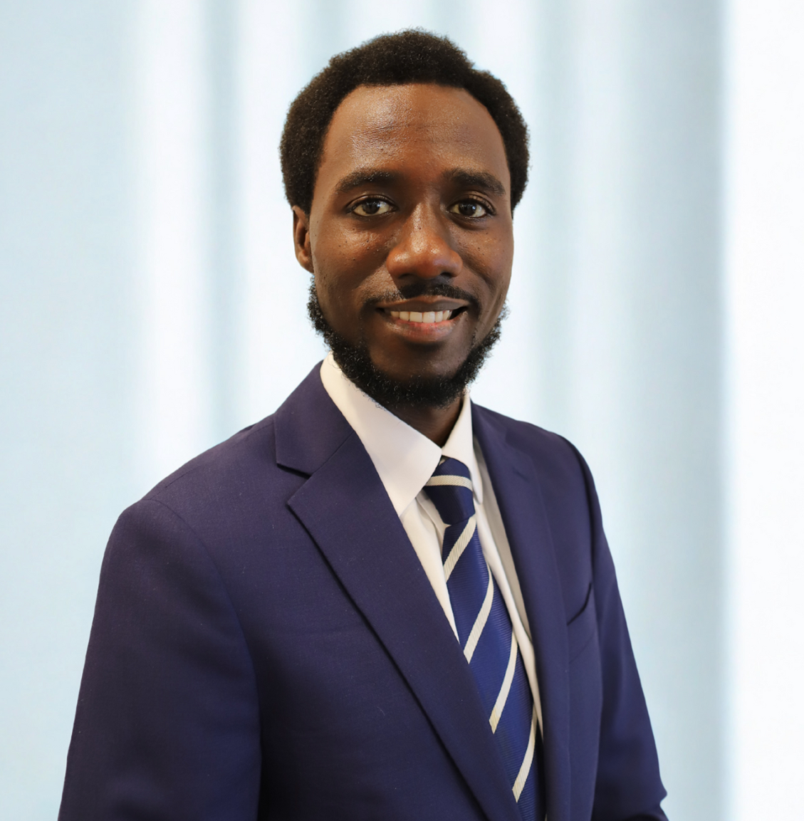 Oludolapo Akinkugbe, Esq., Music Lawyer and Senior Counsel at Sound Logic:  dark hair, wearing a navy blue suit, white shirt, and striped tie, smiling in front of a light blue background.