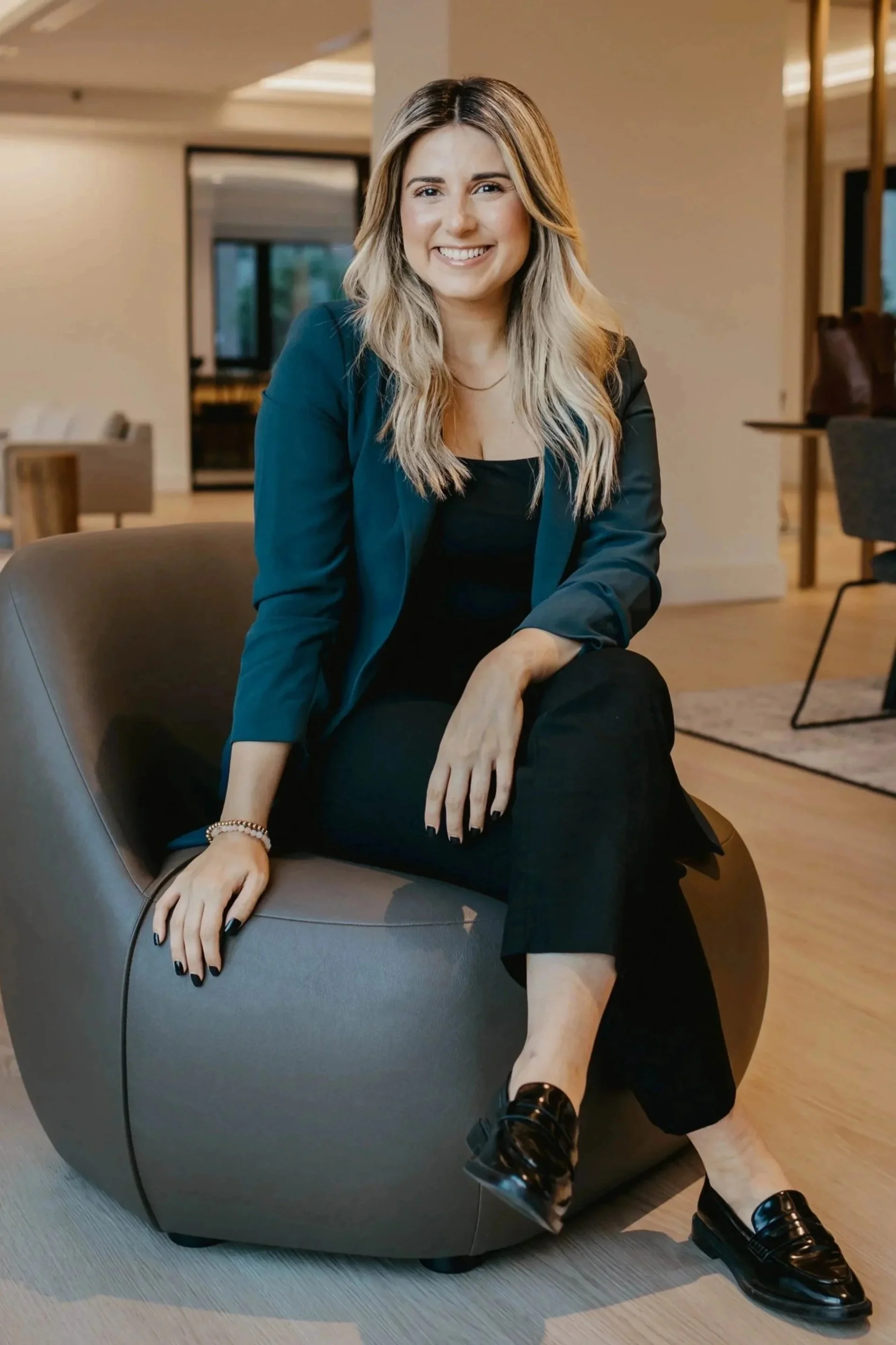 Lisandra Aguiar, Esq., Music Lawyer and Junior Counsel at Sound Logic:  blonde hair, wearing a blue blazer, black top, black pants, and black shiny shoes, sitting on a grey modern chair smiling in a contemporary living space.