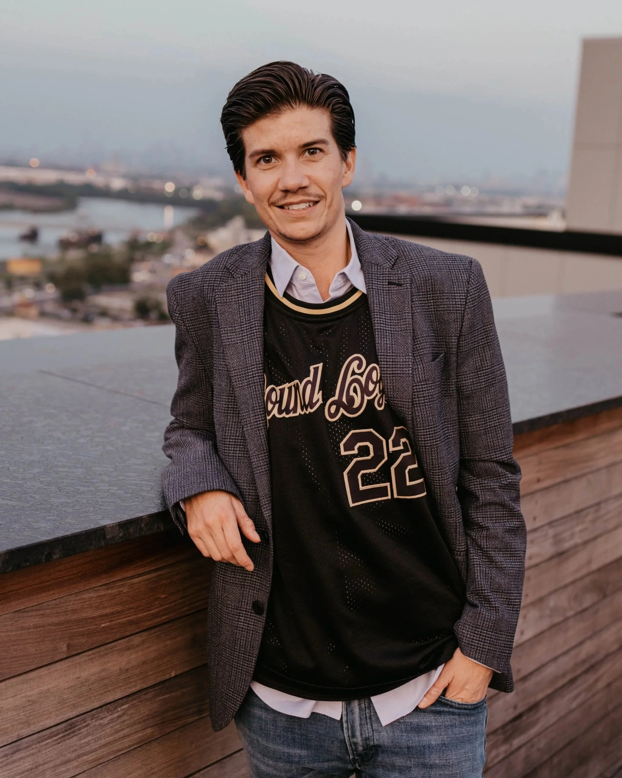 Ashton Monteiro, Esq. entertainment attorney and founder of Sound Logic: smiling while leaning against a rooftop railing, wearing a checked blazer over a black sports jersey and casual jeans, with a cityscape and river in the background during sunset