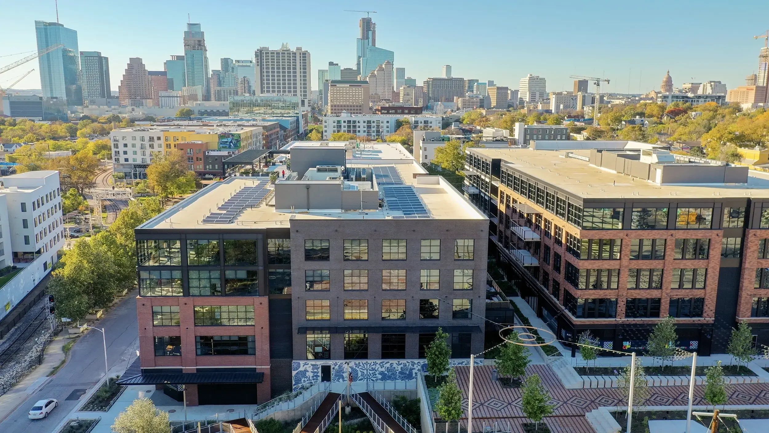 Drone view of Centro, East Austin, TX with the Austin skyline beyond. America Martin's "Armadillo's Song" Otomi mosaic mural visible in the courtyard below.
