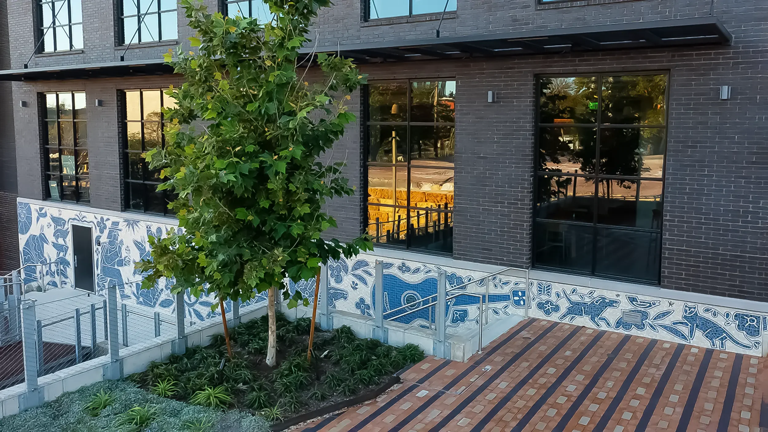 Courtyard view of the left section of America Martin's "Armadillo's Song" at Centro, East Austin, TX. The Colombian-American artist's hand-cut Otomi mosaic mural in blue and white depicts indigenous figurative forms, animals, and botanical motifs alo