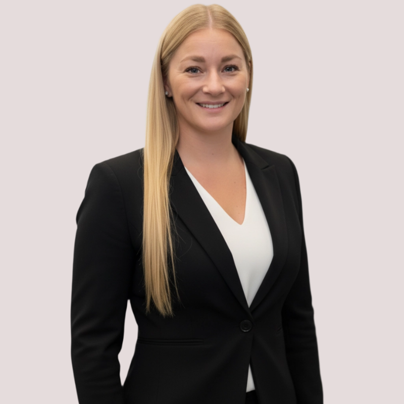 A woman with long blonde hair, wearing a black blazer and a white top, smiling at the camera against a plain light background.