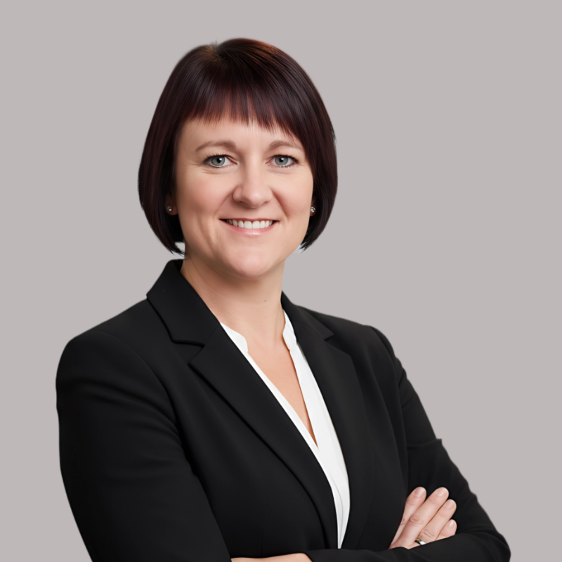 A professional woman with short dark red hair, wearing a black blazer and white blouse, smiling with arms crossed against a gray background.