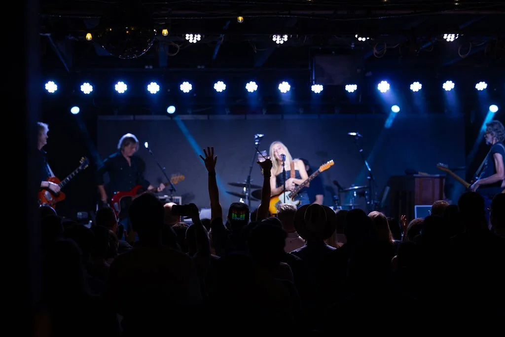 A live band performing under stage lighting with an audience in front.