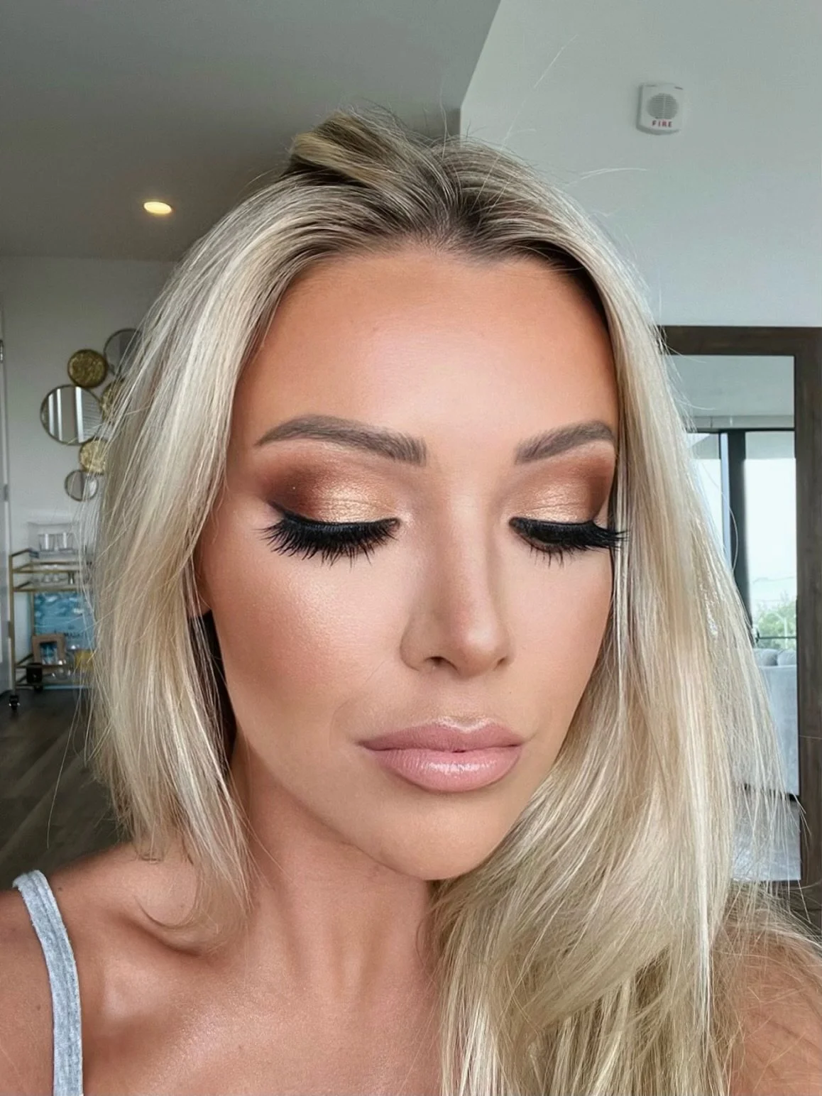 A woman with blonde hair and makeup with bronze eyeshadow, black eyeliner, and long eyelashes, closed eyes, in an indoor setting.