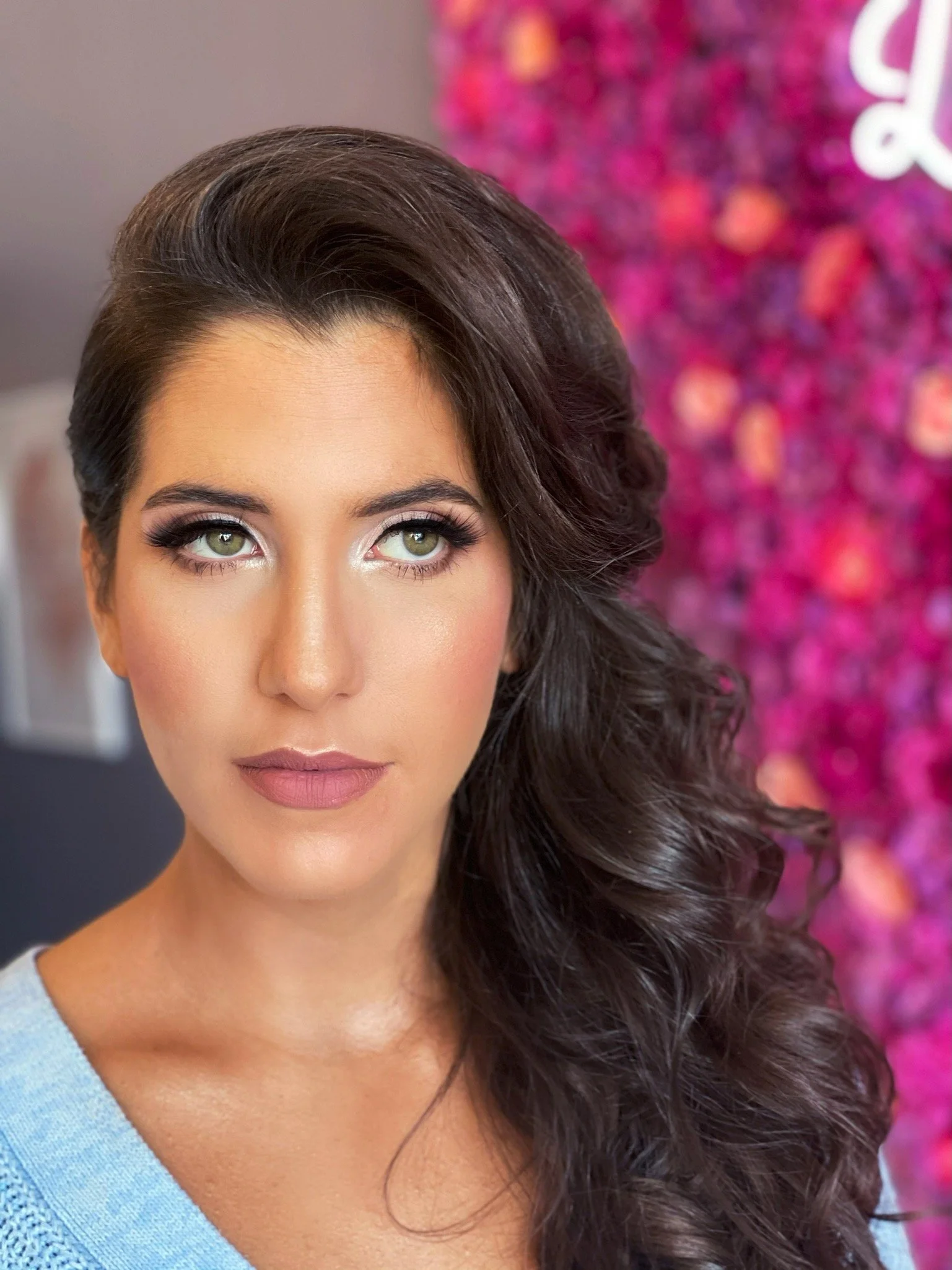 Portrait of a woman with long, dark, wavy hair and makeup, standing in front of a pink and purple floral background.