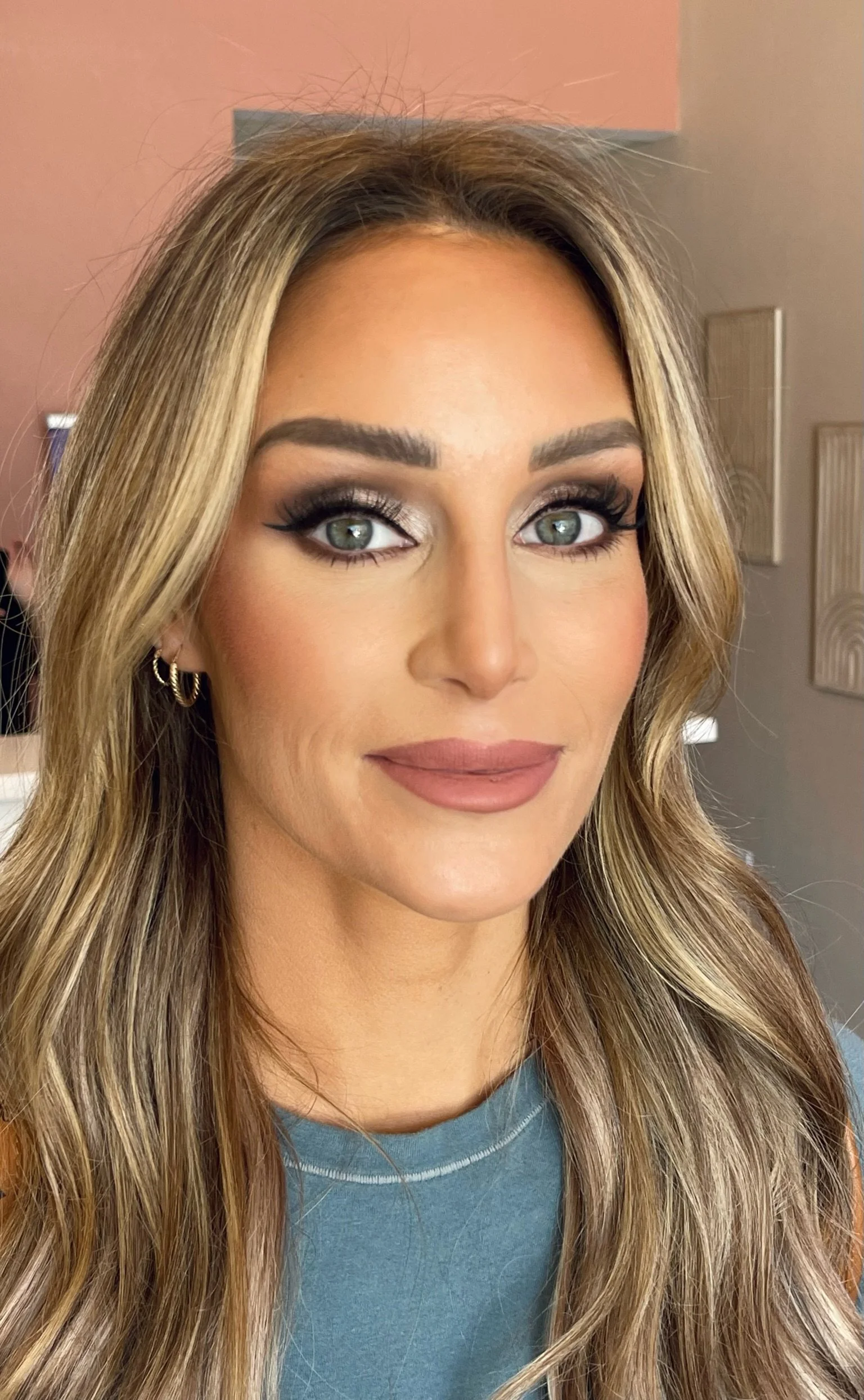 Close-up of a woman with long wavy blonde hair, makeup including silver eyeshadow, winged eyeliner, and matte nude lipstick, wearing small gold hoop earrings and a blue top.