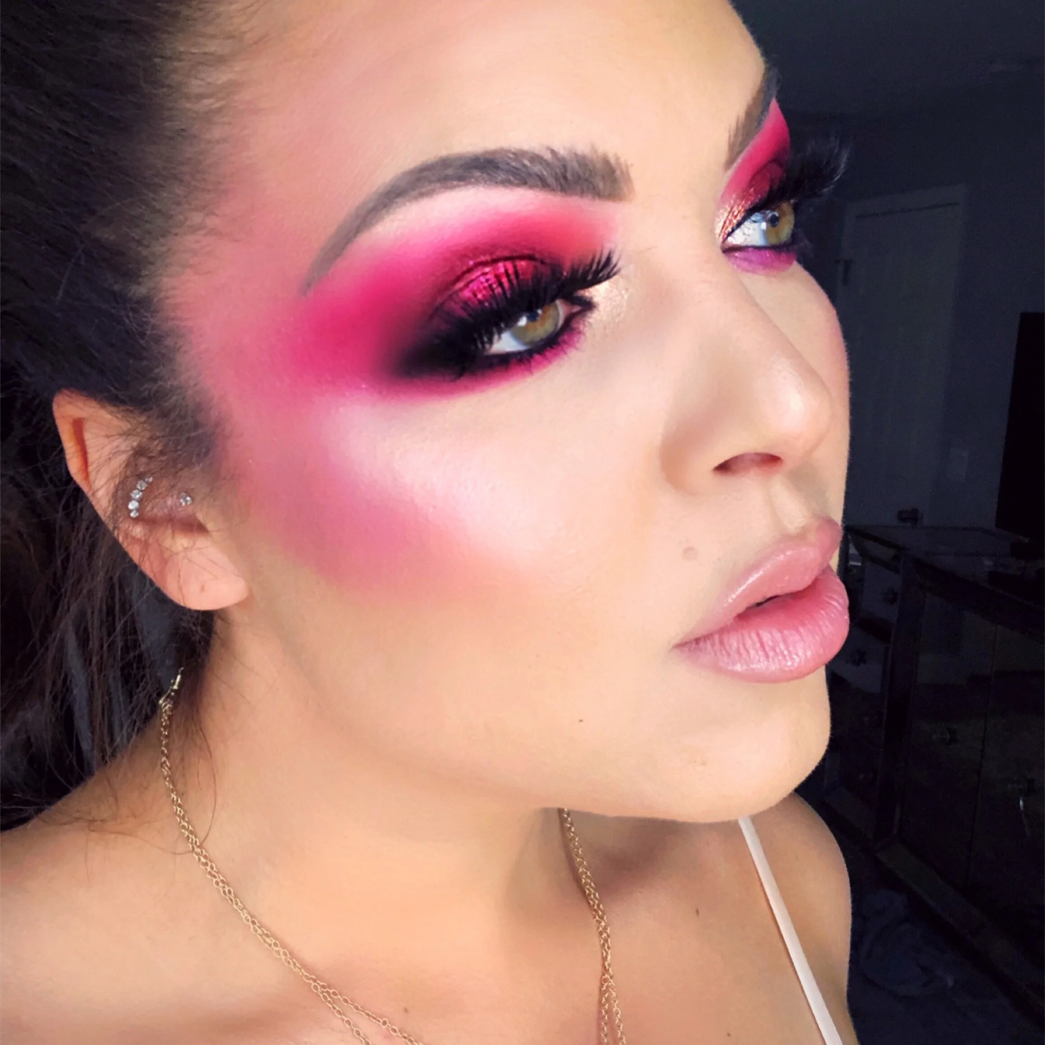 Close-up of a woman's face with dramatic pink and black eye makeup, long false eyelashes, glossy lips, and gold jewelry.