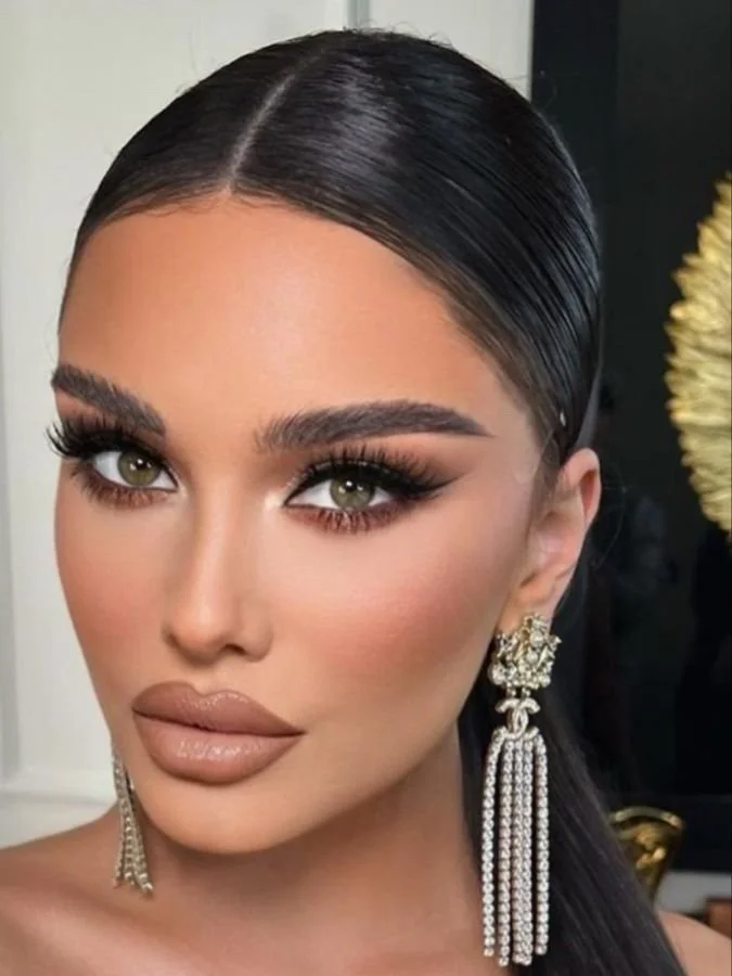 A woman with styled dark hair, makeup with bold eyelashes and contour, wearing large ornate earrings with rhinestones.