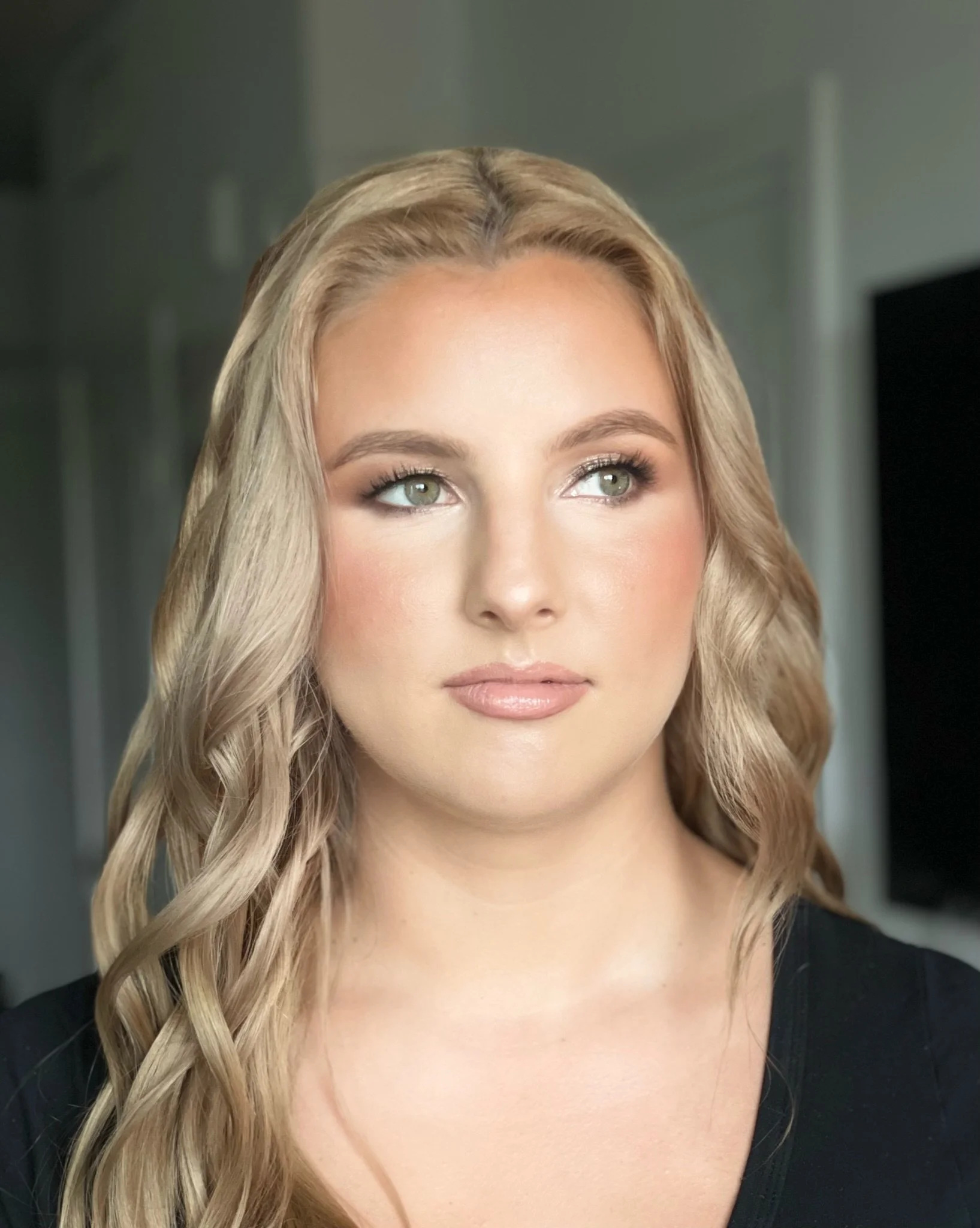A woman with long blonde wavy hair and fair skin looking off to the side, wearing makeup with a natural look, against a blurred indoor background.