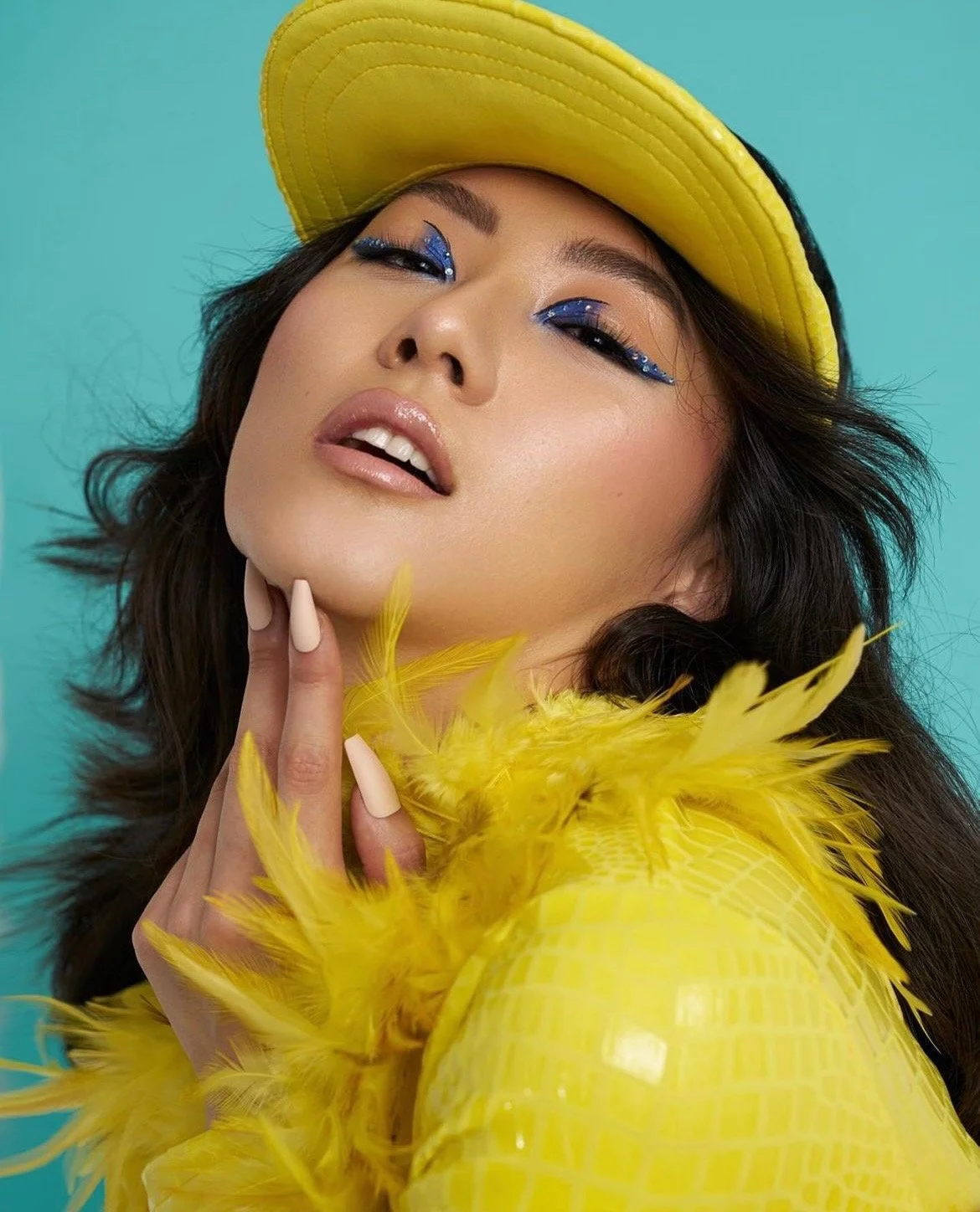 A woman wearing a yellow hat and feathered yellow fashion with striking blue eye makeup, posing against a teal background.