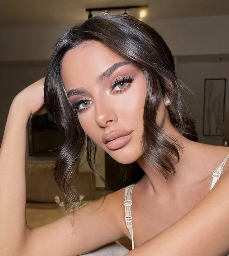 A young woman with shoulder-length wavy dark hair, makeup, and a neutral expression, resting her head on her hand in a well-lit indoor setting.