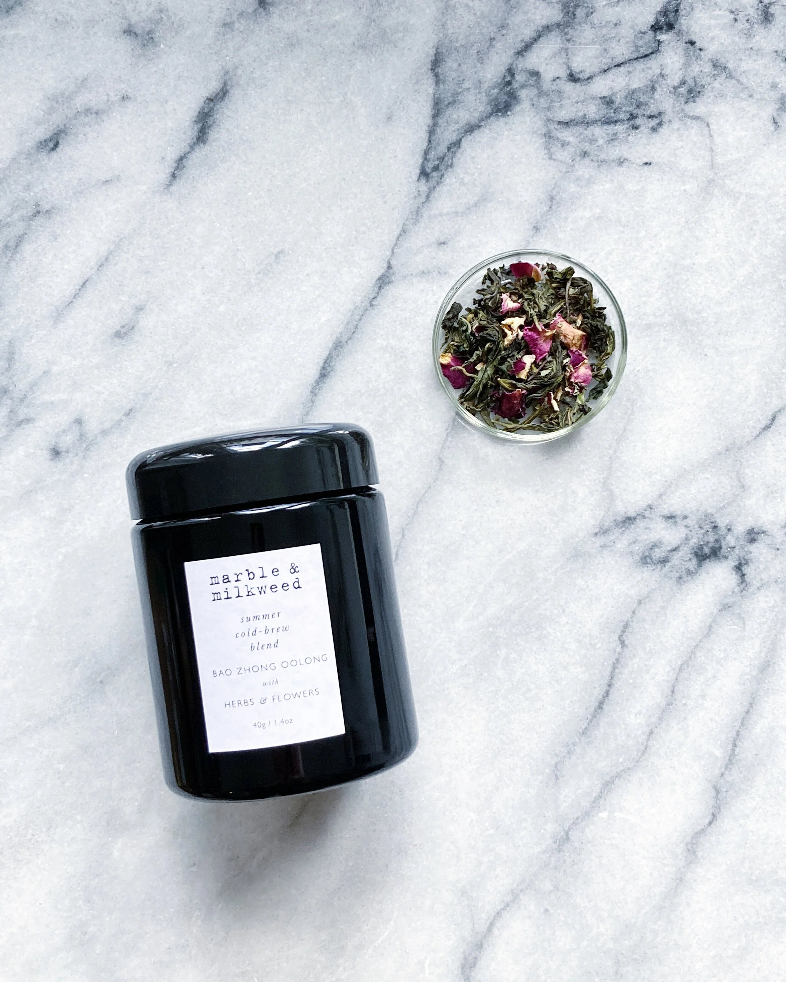 seen from above on a white marble surface, a clear round glass dish holds organic green oolong tea leaves with pink rose petals, alongside is a black glass jar of tea with white label & black text