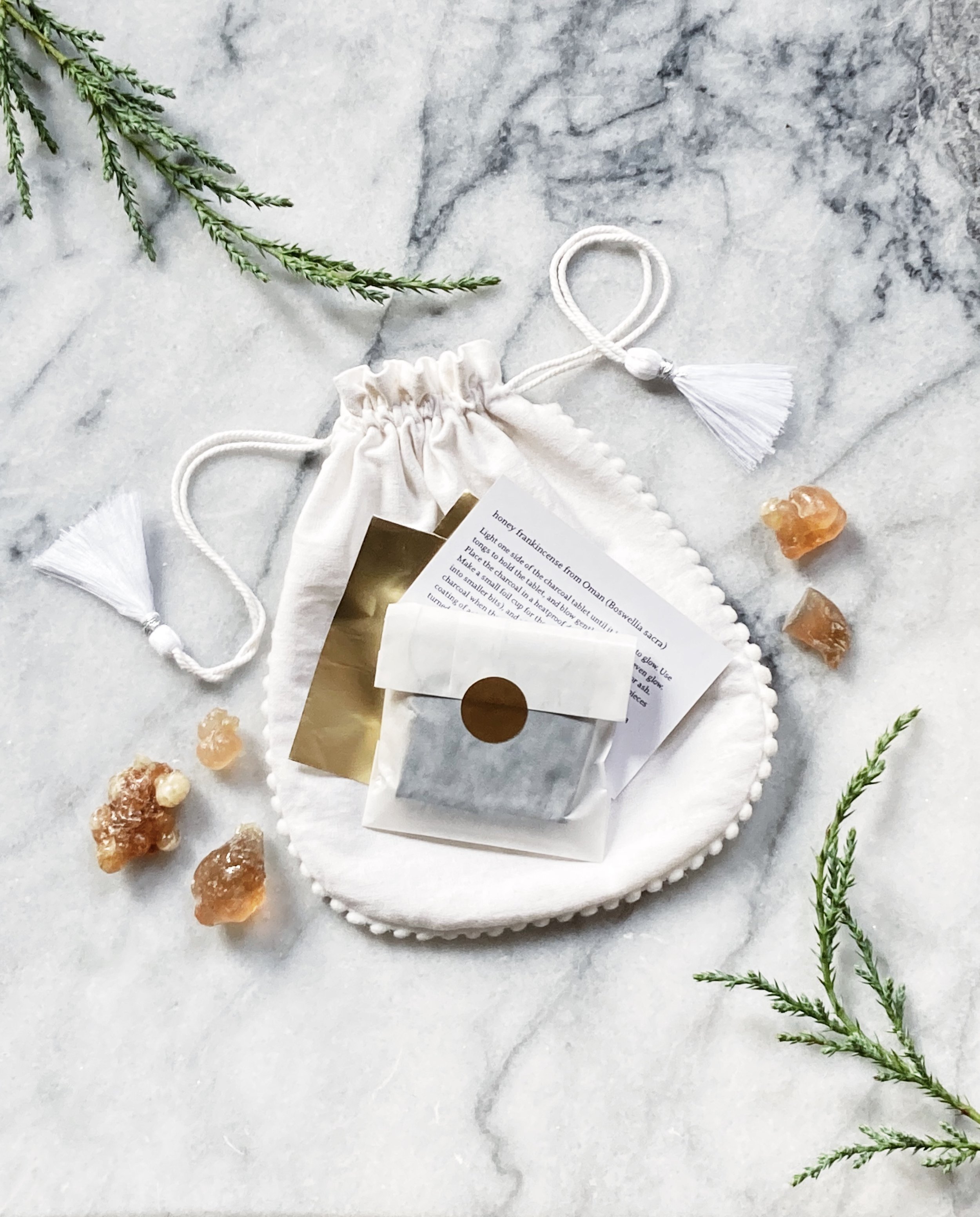 seen from above on a white marble surface, a white drawstring gift bag holding golden frankincense resin, a white envelope holding incense charcoal, gold foils, an instruction card and juniper sprigs