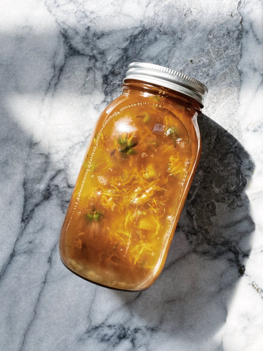 a glass jar full of golden calendula infused sunflower oil sits diagonally on a marble surface in the sunlight