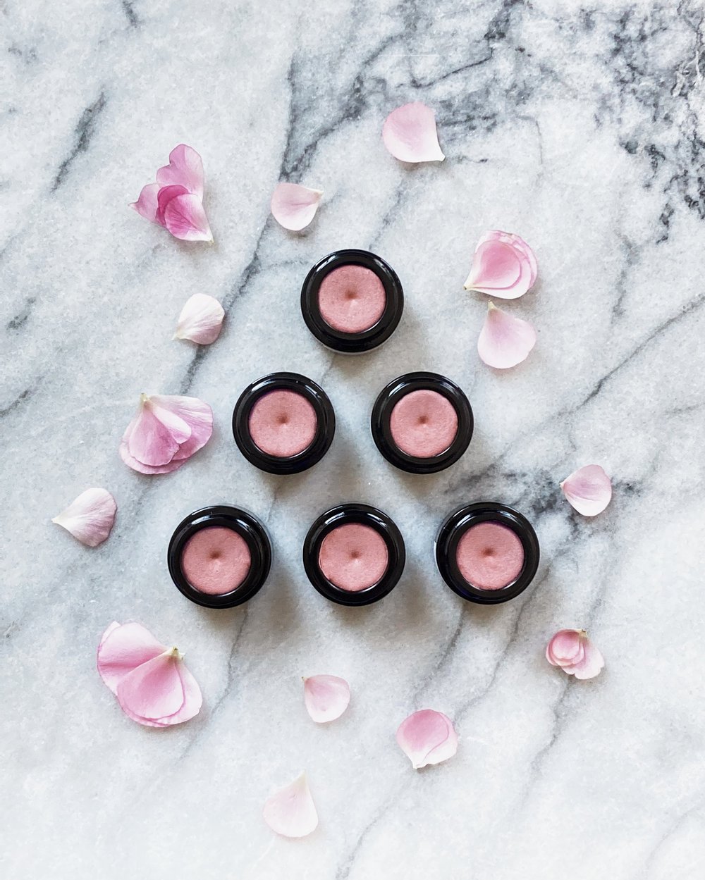 small black jars of pink luminescent shimmer balm arranged in a triangle together with pink flower petals on a white marble surface