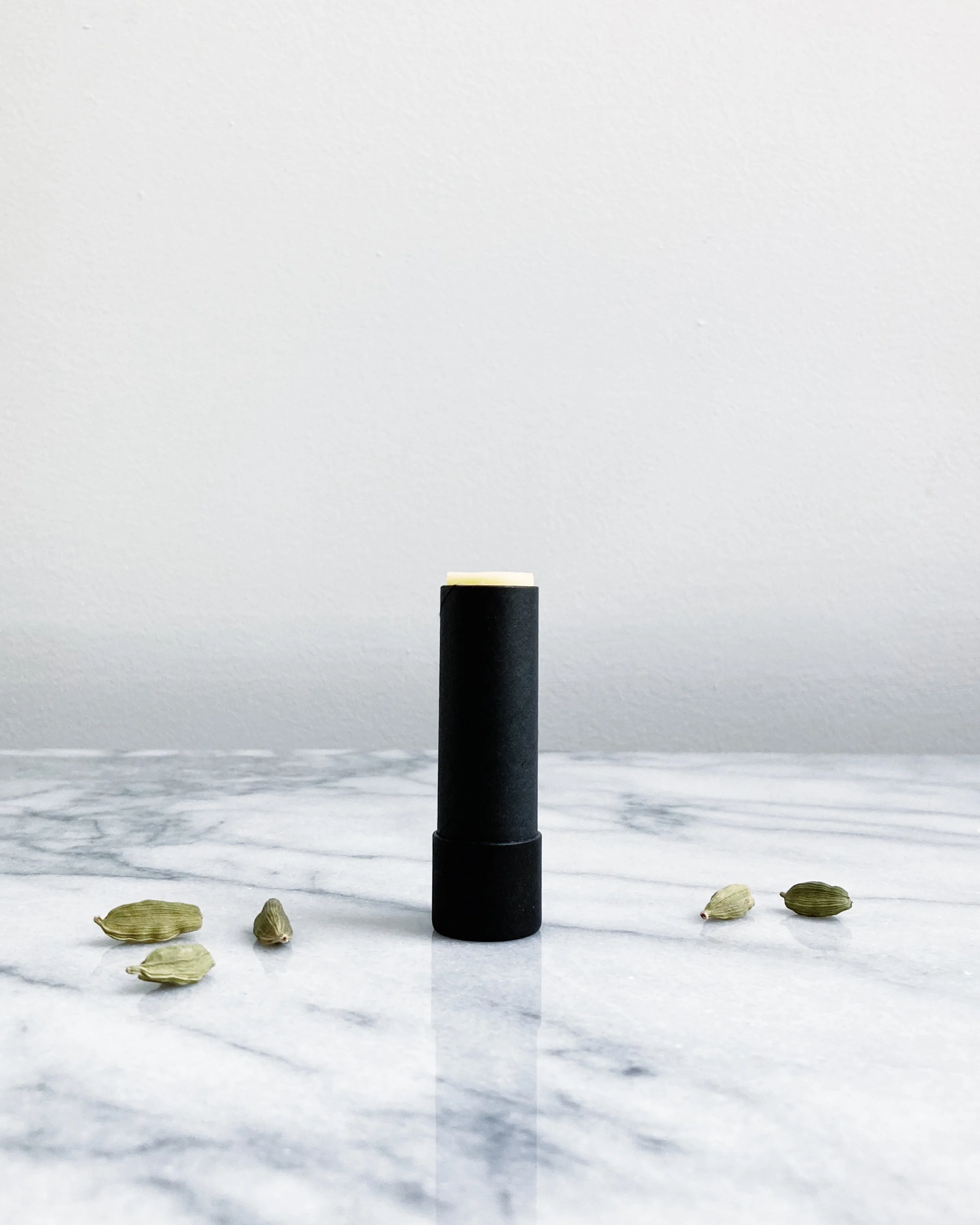 Lip balm tube with cardamom pods scattered around on a marble surface against a white wall.