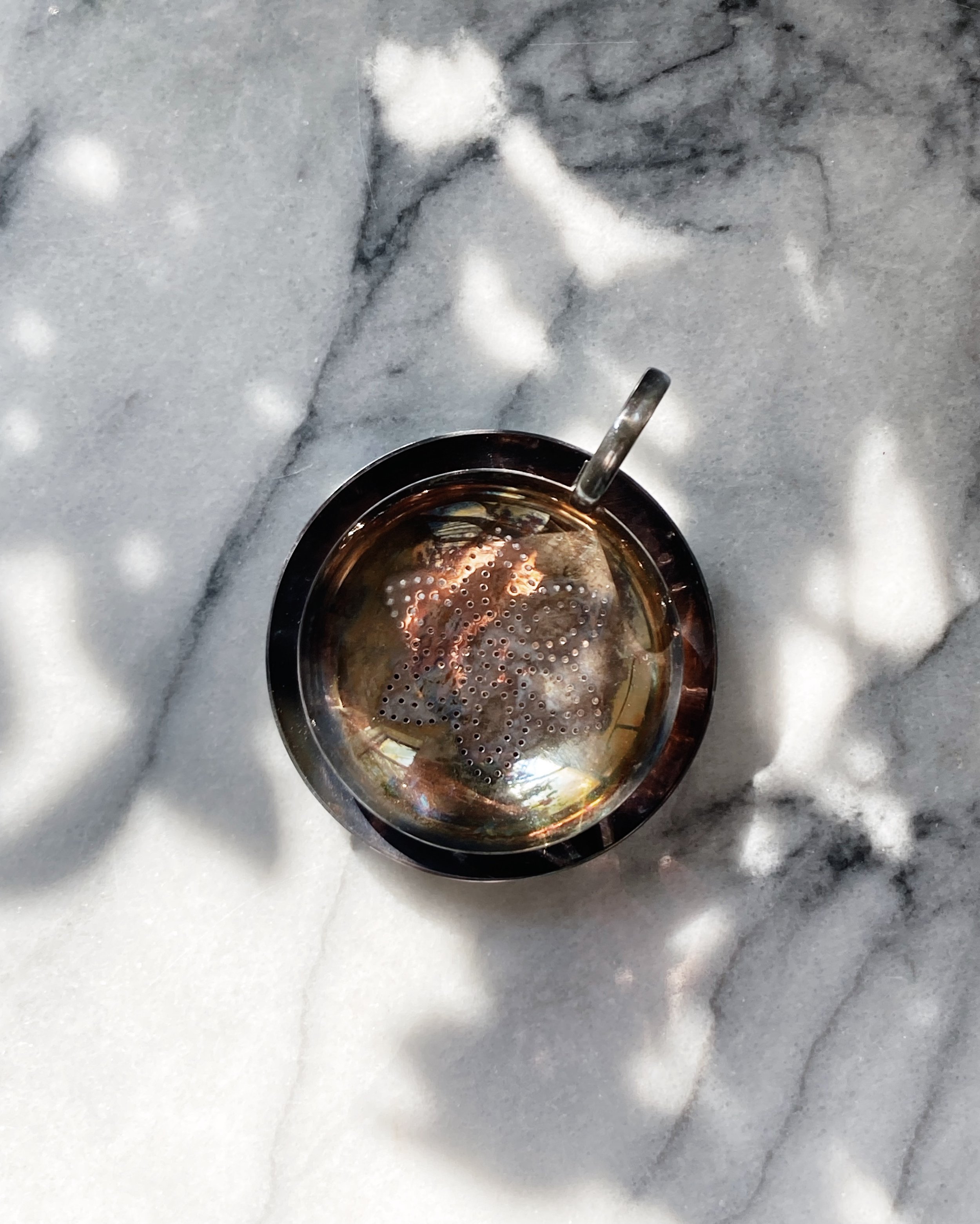 A vintage tea strainer with an ornate perforated bowl showing colorful patina sits in a matching tray with decorative feet, displayed on white marble surface.