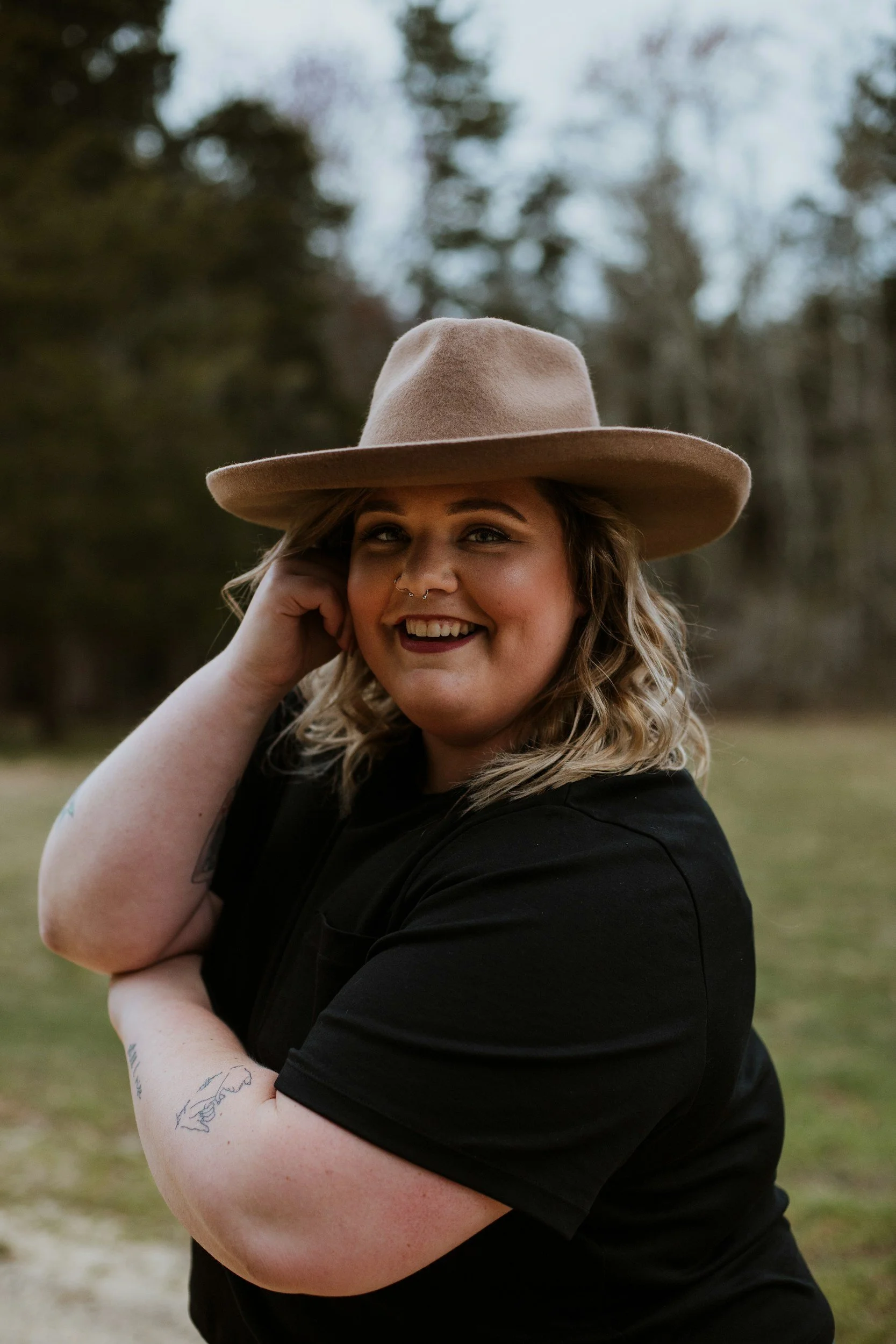 A woman smiling outdoors, wearing a large tan hat and black shirt, with curly blonde hair and tattoos on her arms, standing in a grassy area with trees in the background.