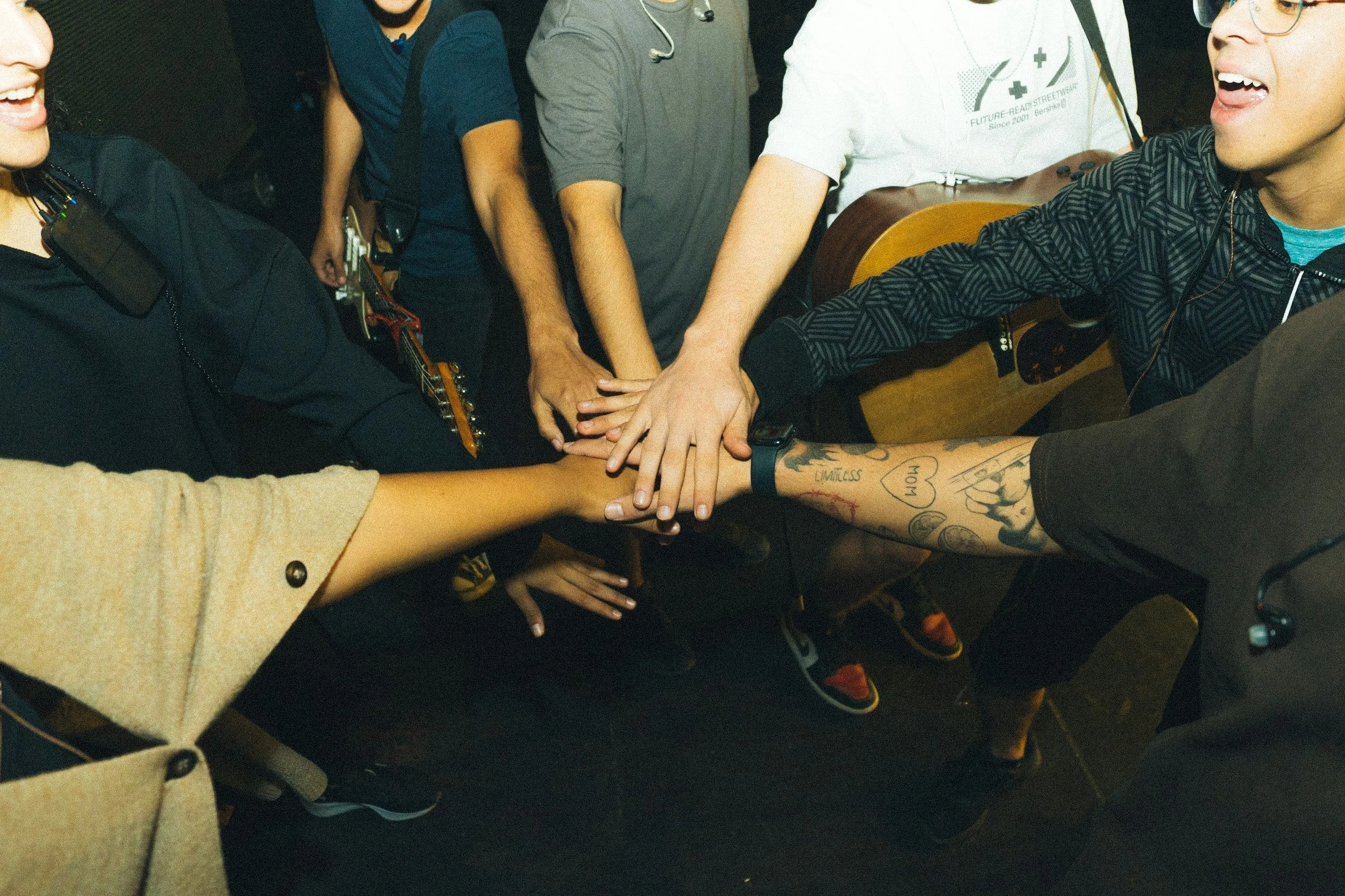 Group of diverse people placing their hands together in a circle, celebrating or showing unity, with some holding musical instruments like a guitar.