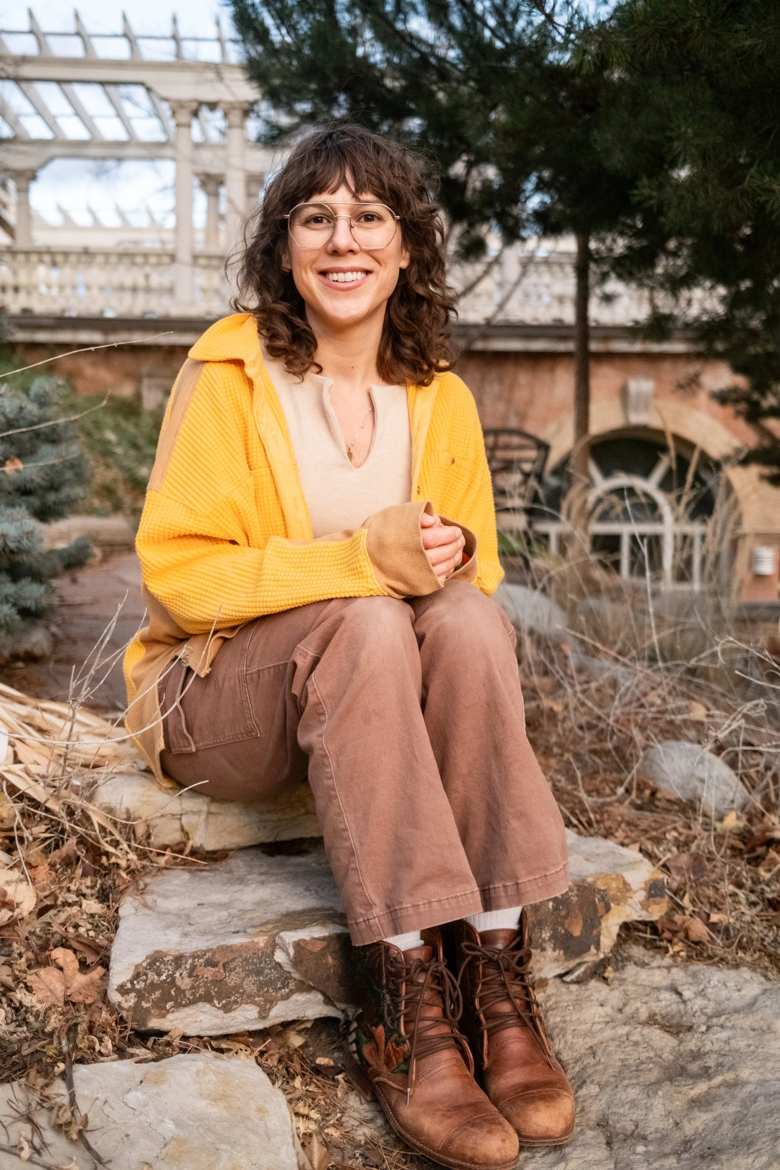 A woman with curly brown hair, glasses, and a bright yellow jacket, sitting outdoors on a stone step, smiling at the camera. She is wearing brown pants and brown lace-up boots, with a background of trees, a wooden structure, and autumn leaves.