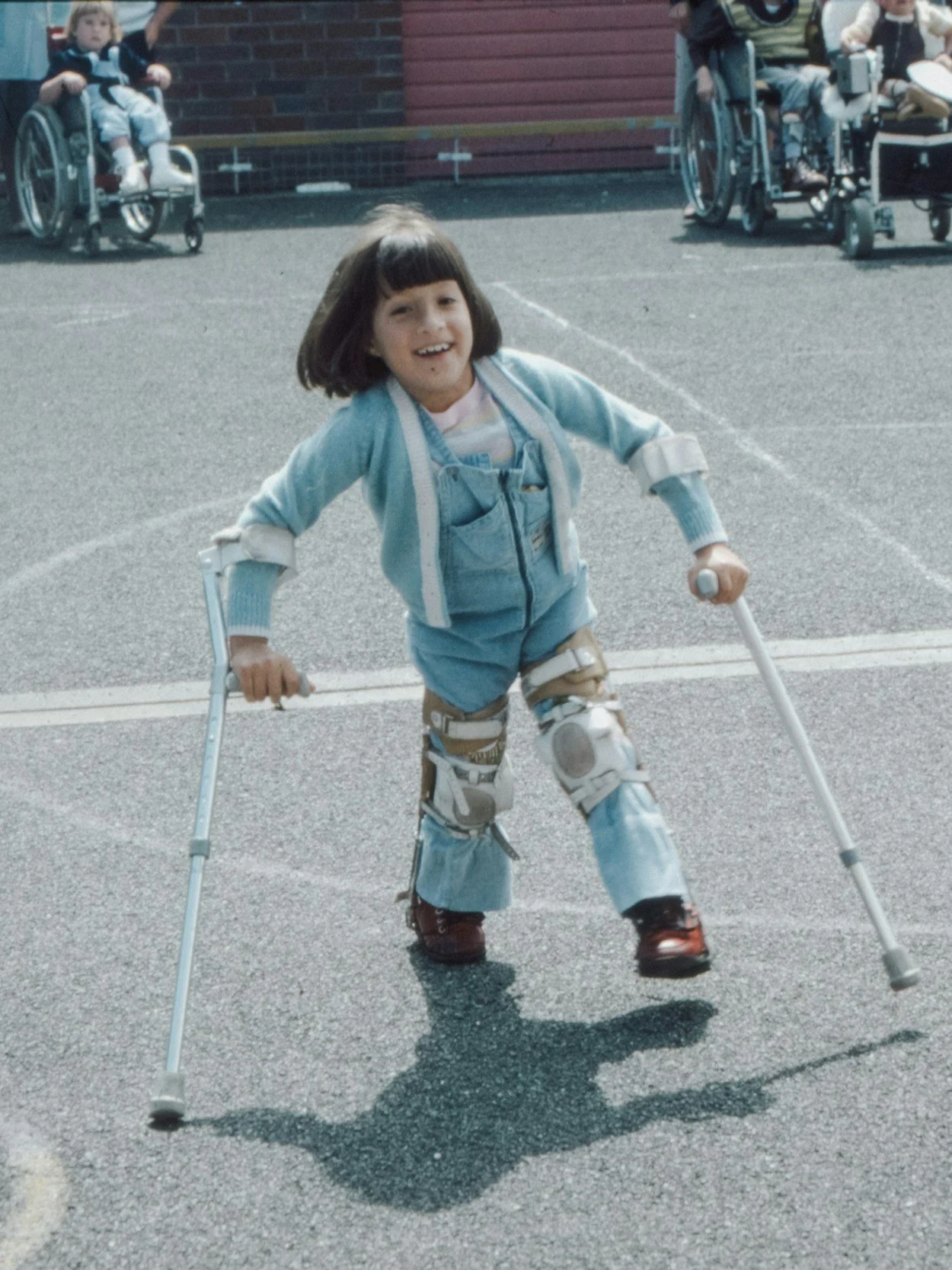 A young girl with a prosthetic leg and crutches standing on a paved outdoor area, smiling and enjoying herself.