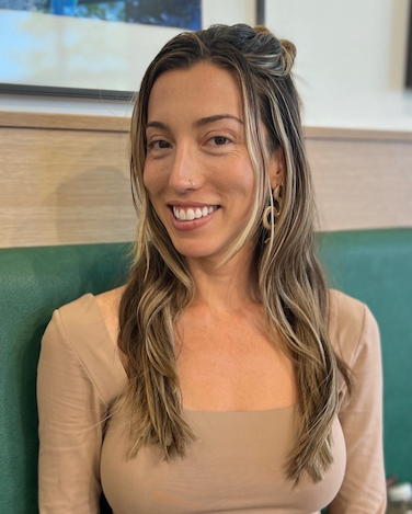 A woman with long wavy hair, smiling, sitting in a restaurant or cafe with a green booth and window behind her.