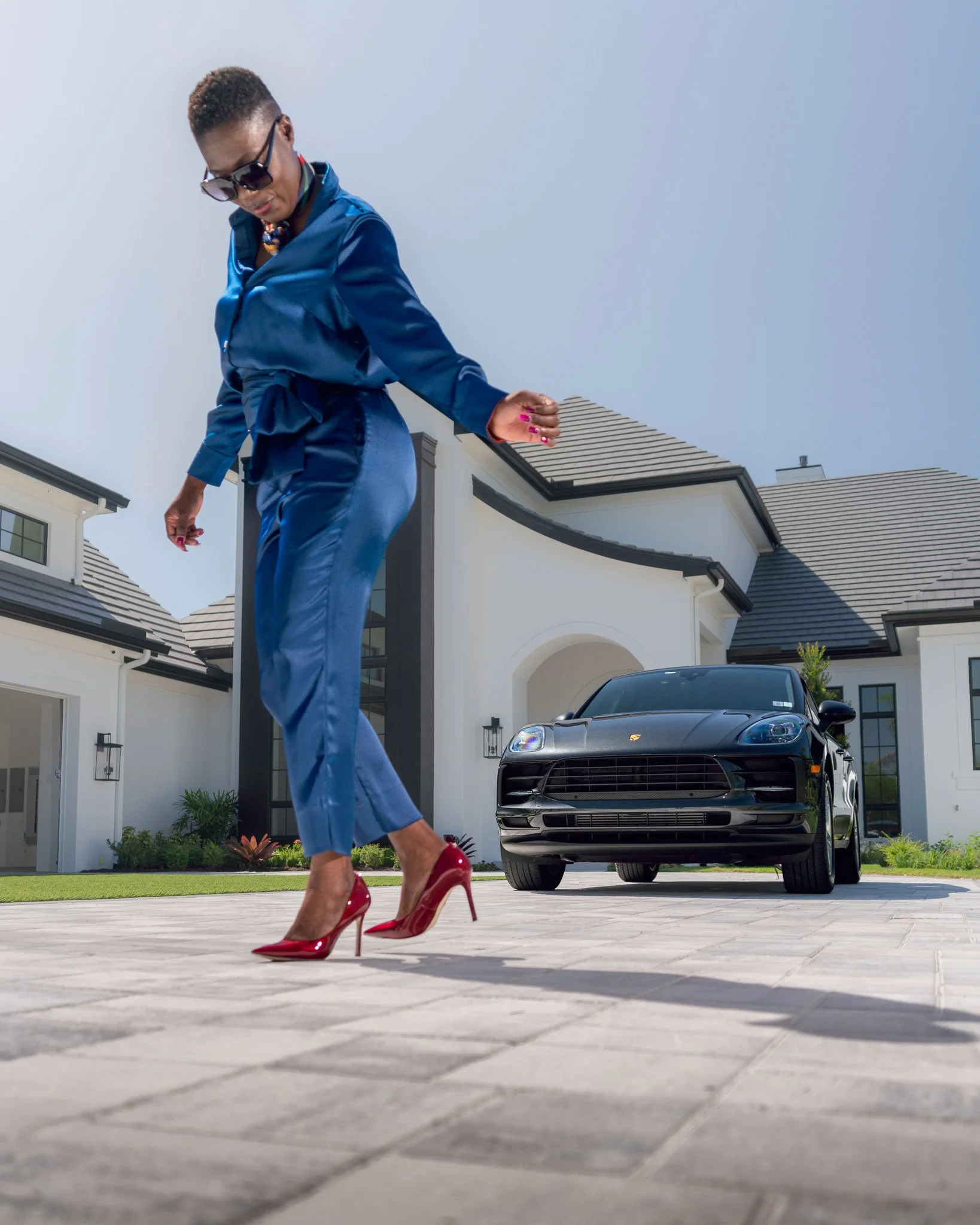 A woman in a shiny blue dress and red high heels walks on a paved driveway in front of a modern white house with a Porsche car parked nearby.