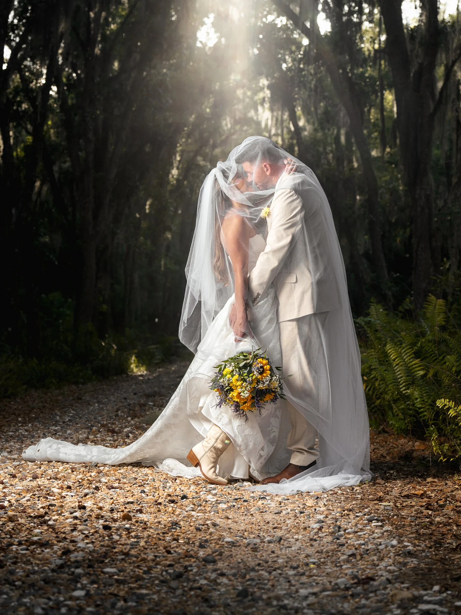 A bride and groom embracing in a wooded outdoor setting, partially covered with sunlight, with the bride holding a bouquet of yellow and purple flowers.