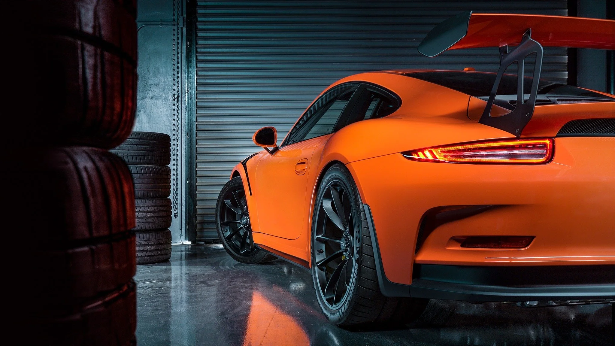 Orange sports car parked inside a garage with tires stacked on the left and a metallic roll-up door in the background.