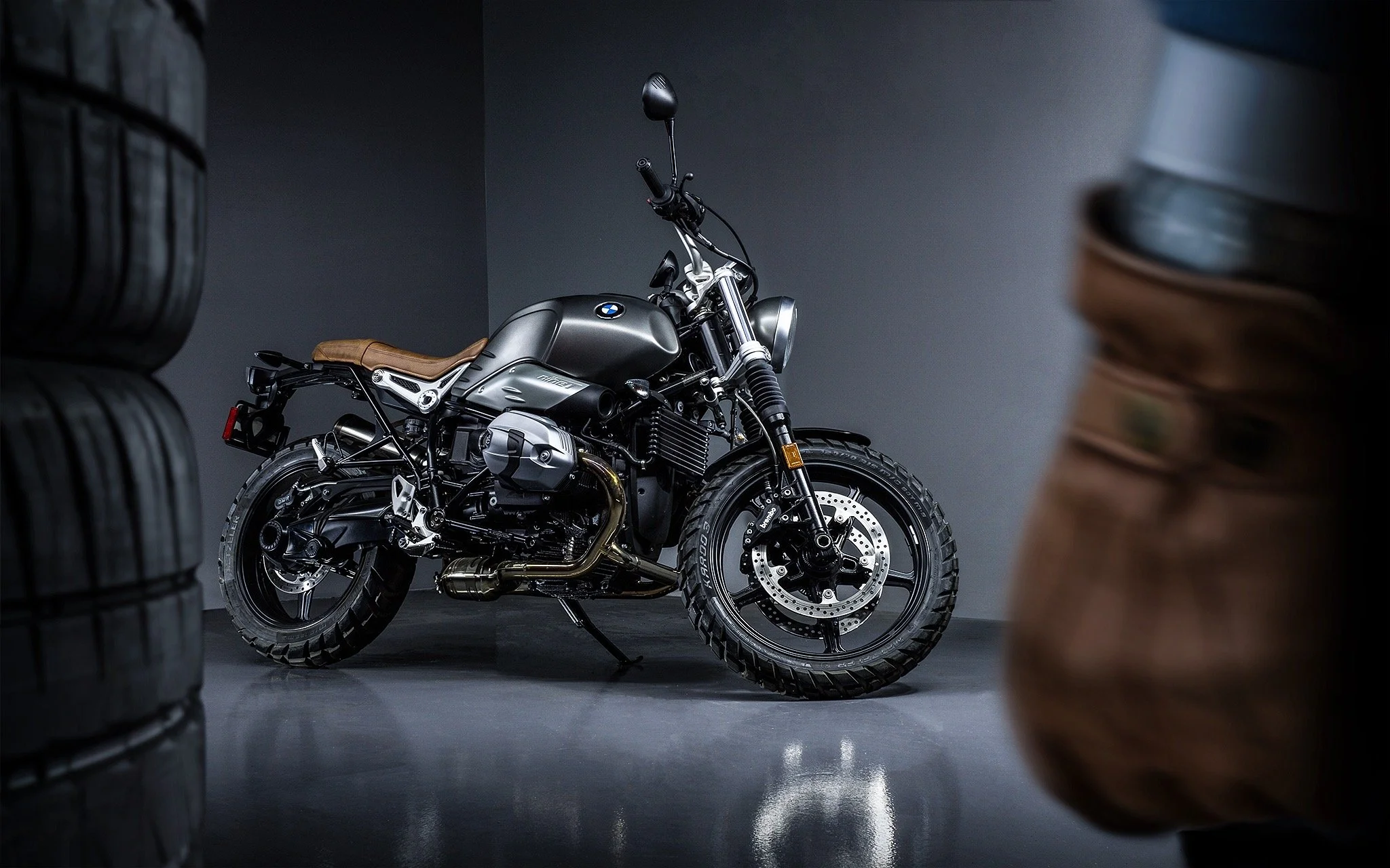 A matte black BMW motorcycle with tan leather seat parked indoors, viewed from the side with tires stacked on both sides of the frame.