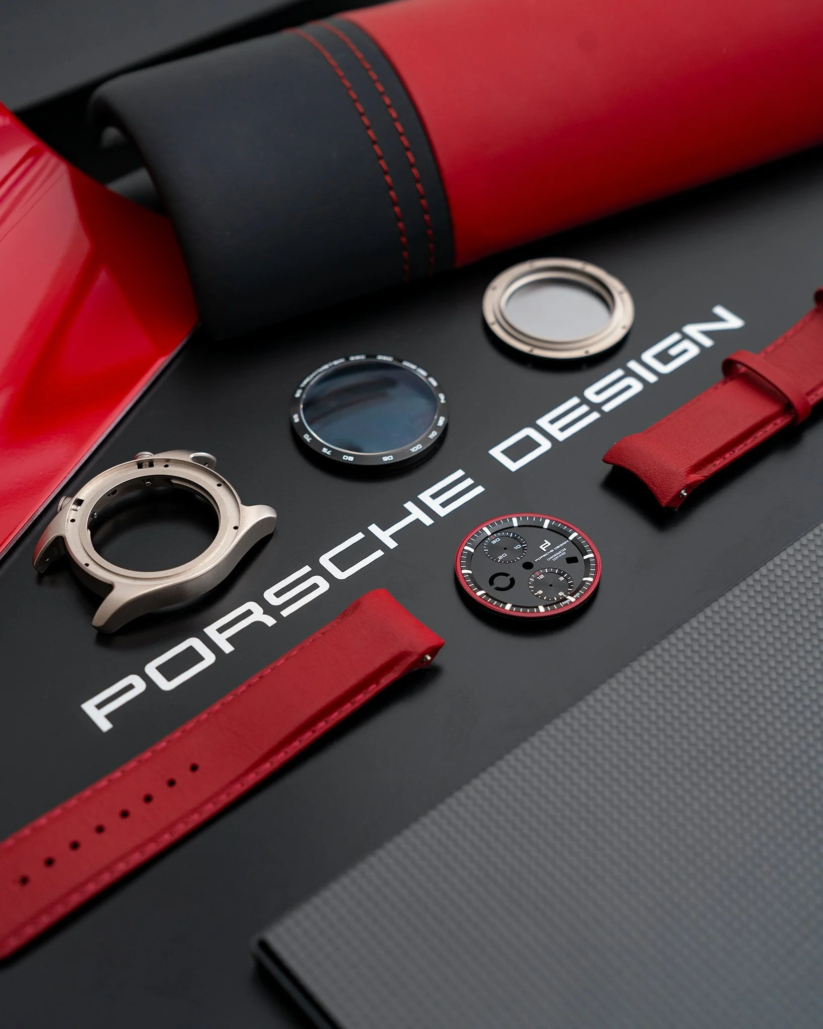Disassembled Porsche watch components on a black surface with 'Porsche Design' logo, including watch case, dial, bezel, and straps, with red accents.