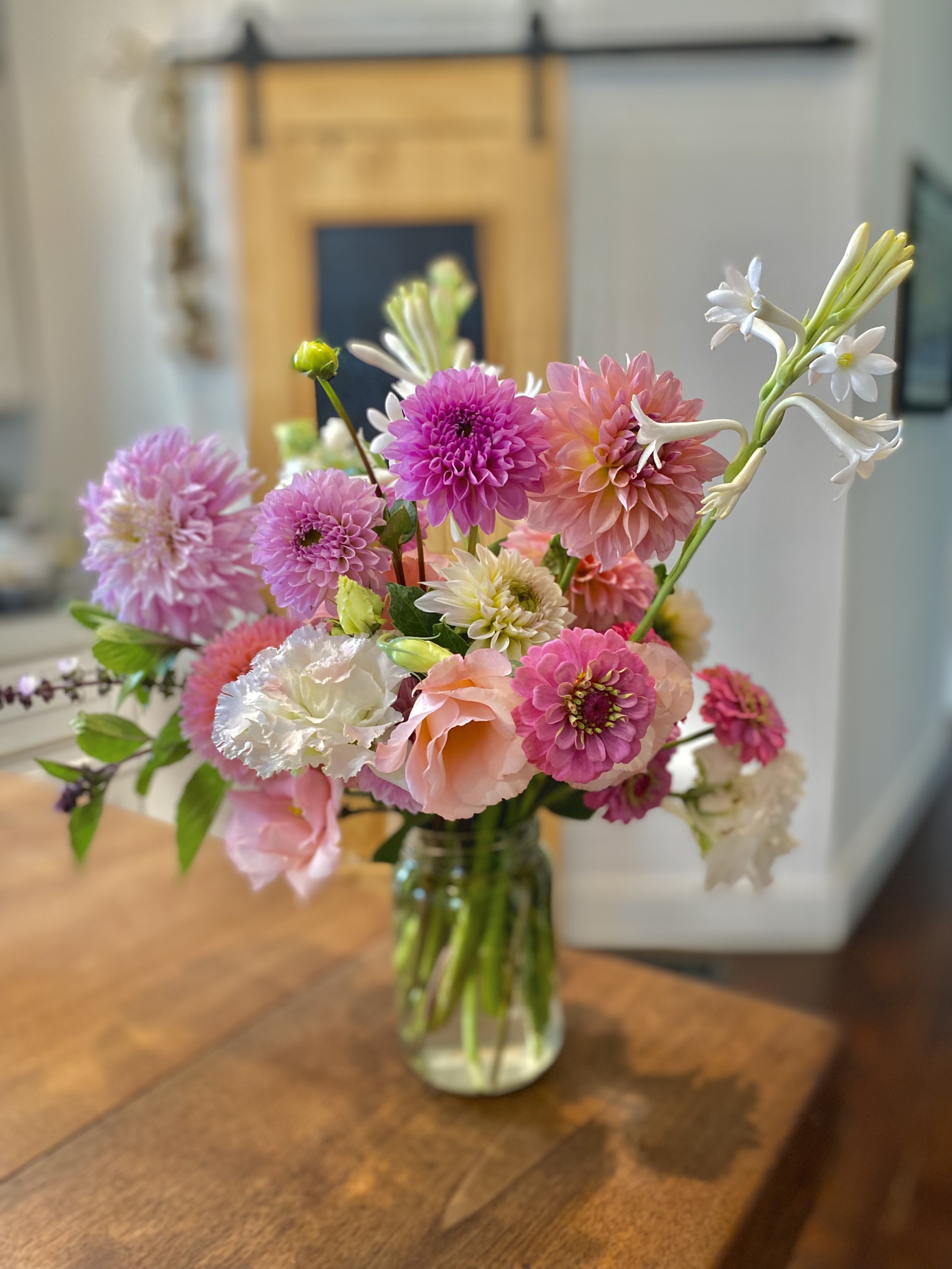 A glass vase with a colorful bouquet of pink, white, peach, and purple flowers on a wooden table, with a blurred background including a wooden fireplace and framed pictures.
